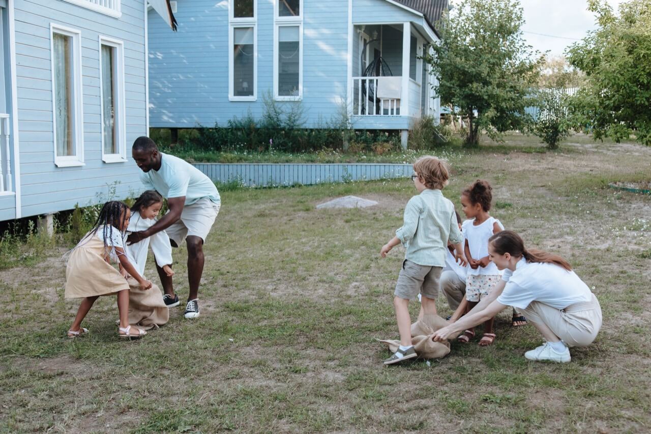 a multiracial family with several children plays a sack race game on the grassy lawn between two light-blue wooden houses on a summer evening.