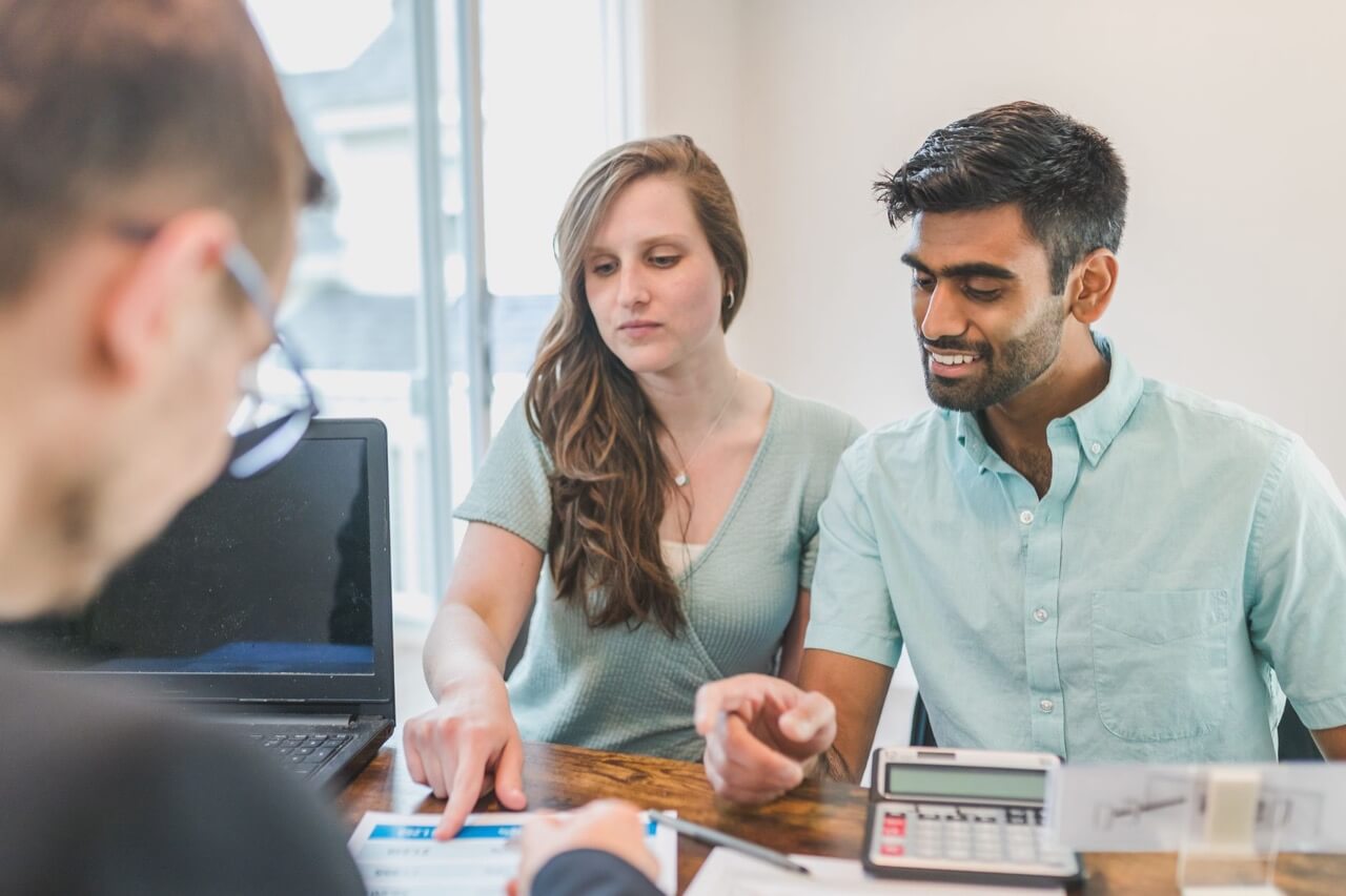 A young couple sits at a desk with a laptop and calculator while a loan officer points out figures on a document.