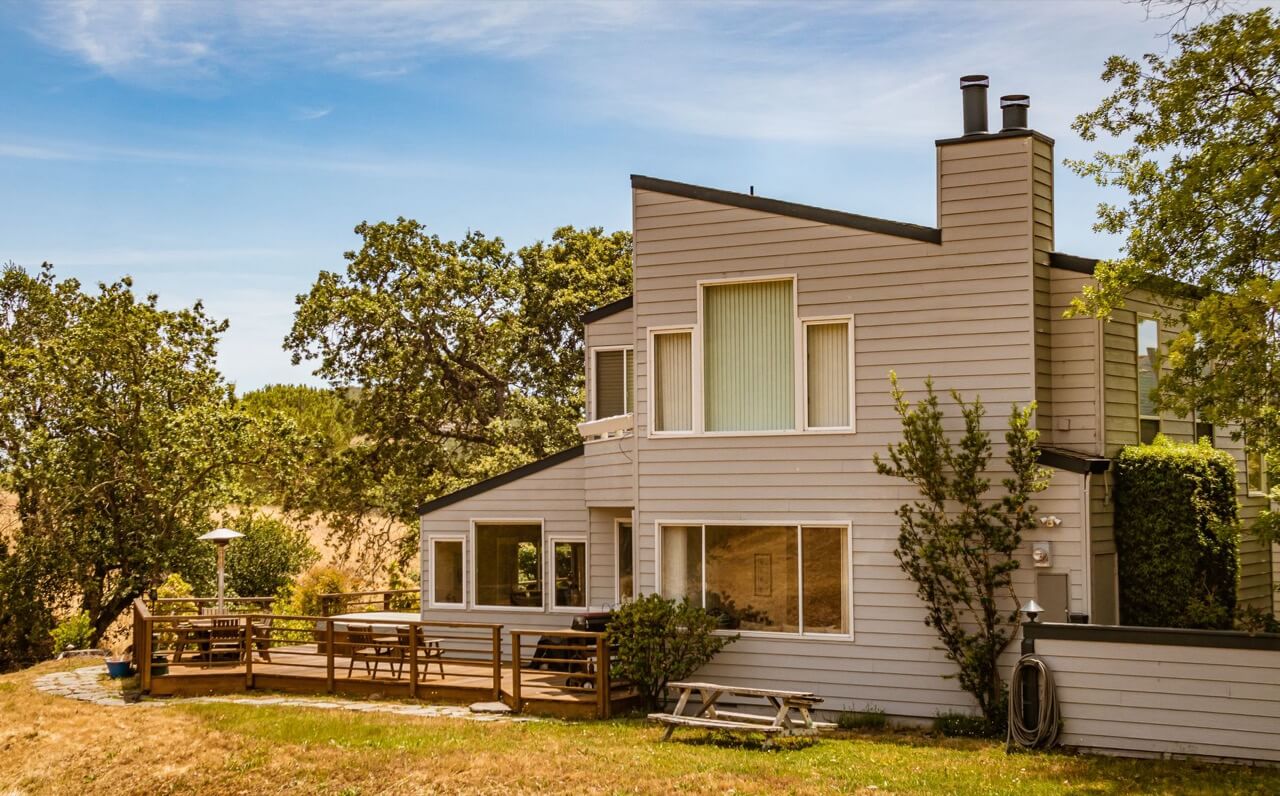 A modern two-story house with light gray siding, a wooden deck, and a backyard under a blue summer sky.