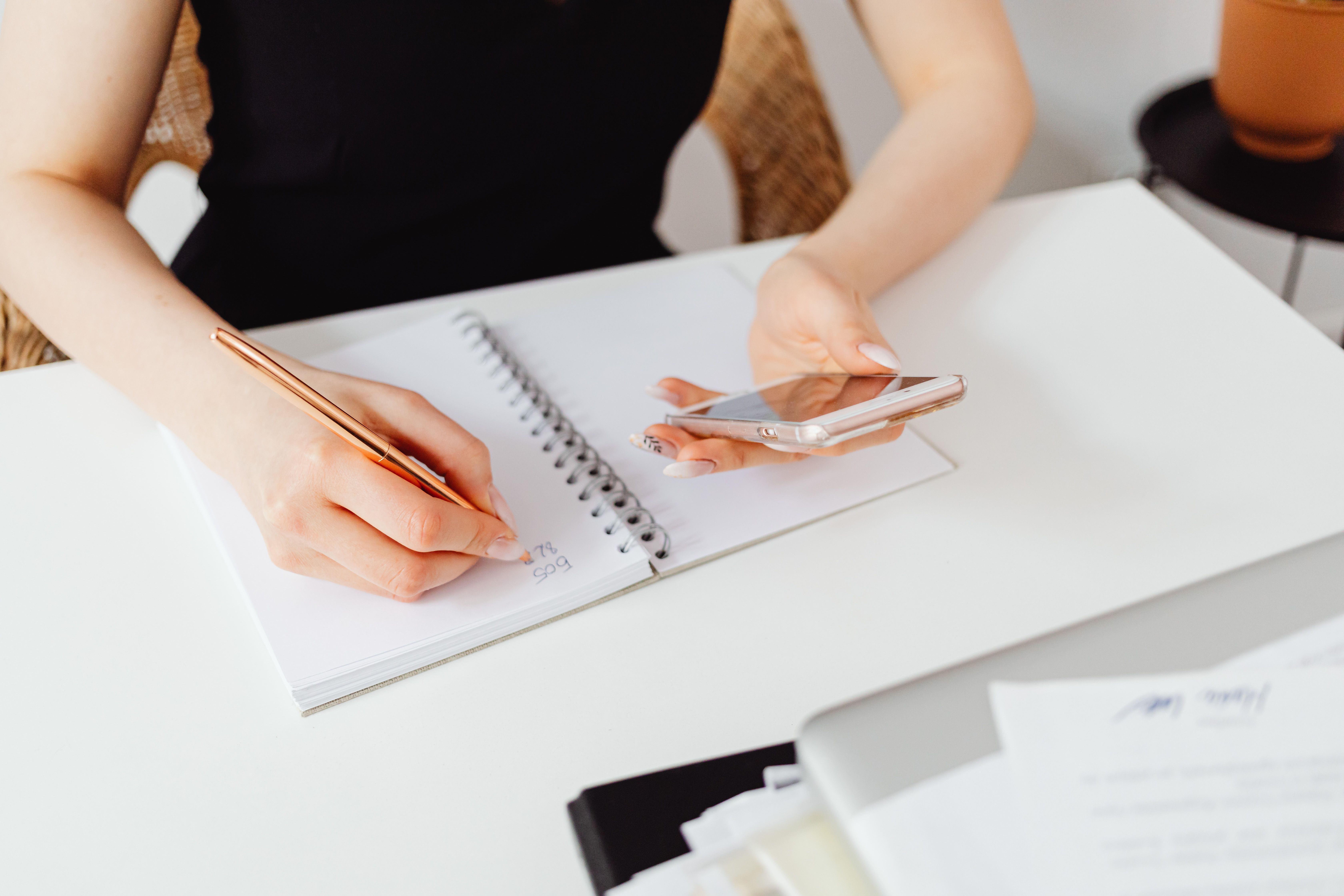 Person holding a smartphone while writing in a notebook.