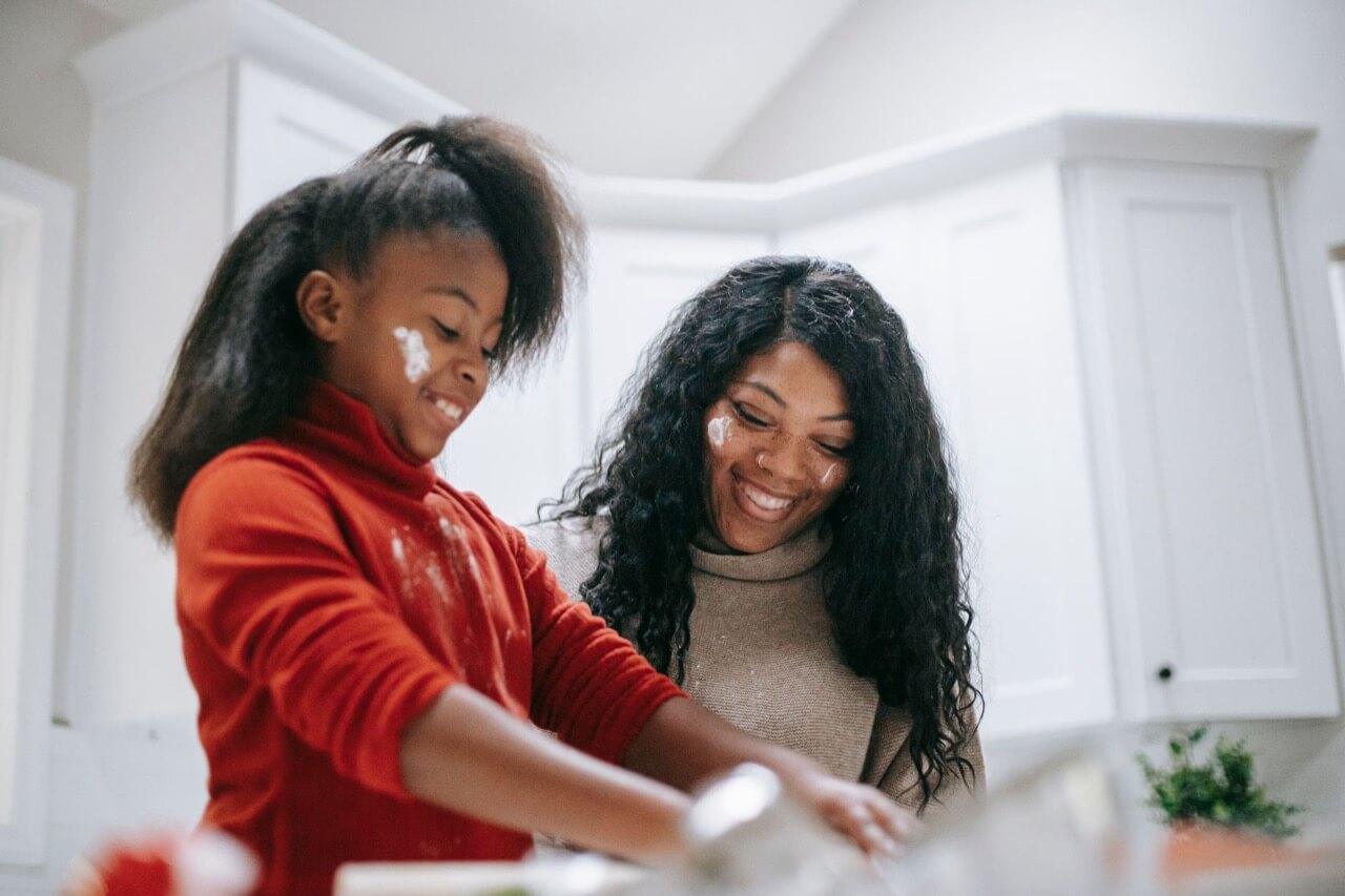 A smiling mother and daughter with flour smudged on their faces bake together at a counter in a bright white kitchen.