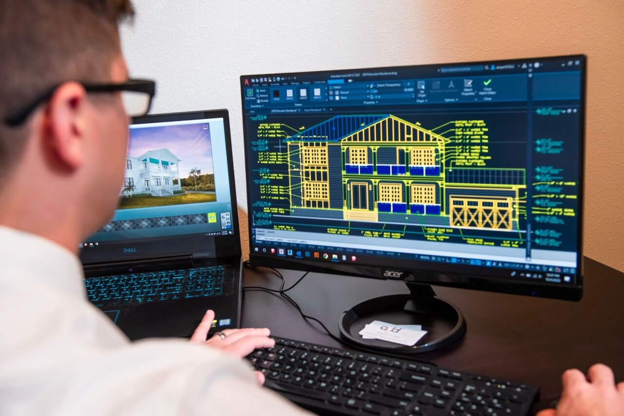 Over-the-shoulder view of a man in glasses and a white shirt working at a dual-monitor workstation, reviewing a residential house elevation drawing in autocad beside a 3d rendering of the home on a laptop.