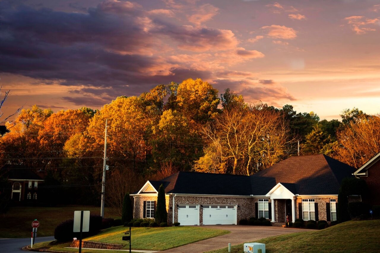 A single-story brick suburban house with a two-car white garage, dark shingle roof and driveway sits on a lawn at sunset