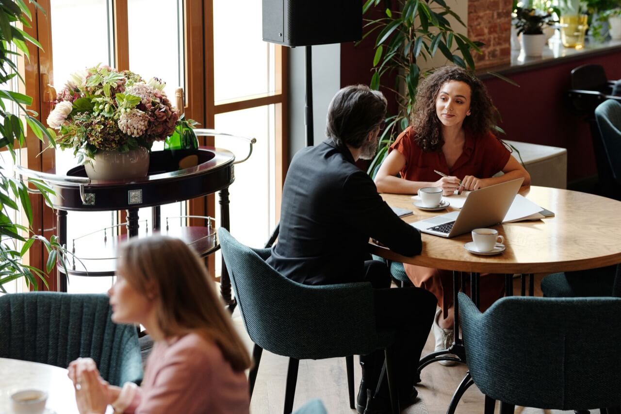 A gray-haired man in a black suit and a curly-haired woman in a rust-red dress review documents beside a laptop at a round cafe table, with a large floral bouquet and another woman drinking coffee in the foreground.
