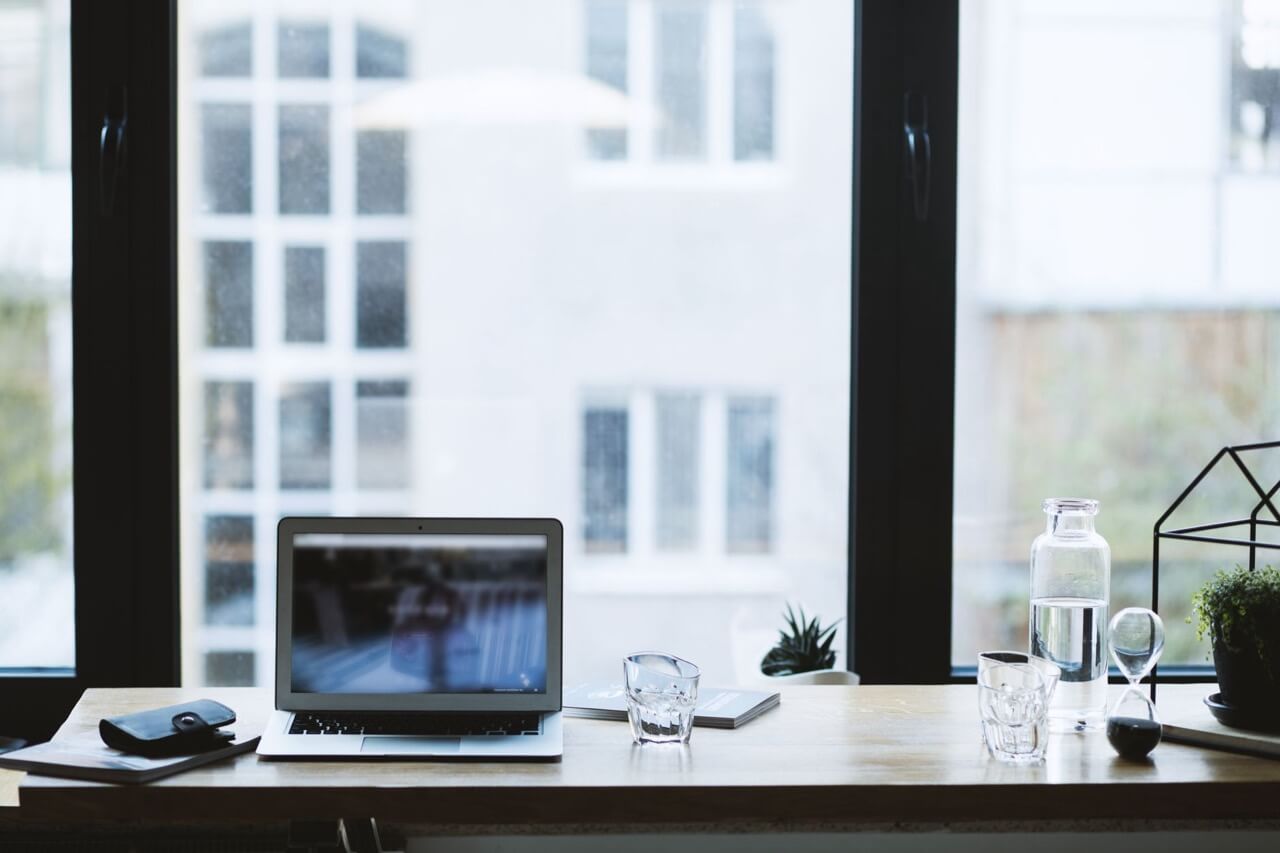 An open laptop sits on a light wooden desk by a large window with blurred white buildings outside, alongside drinking glasses, a water carafe, an hourglass, a wallet, and a small potted plant.