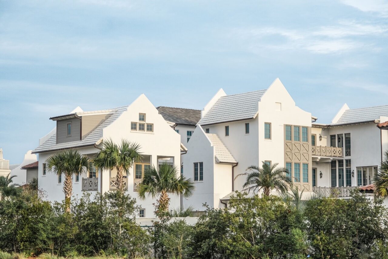 Row of large white stucco luxury homes with steep gabled roofs, gray tile roofing, and wooden balconies, framed by palm trees and green shrubs under a pale blue sky.