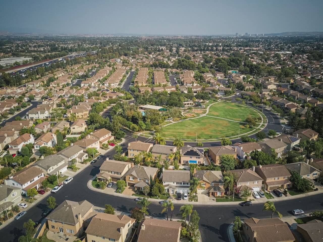 Aerial drone view of a sprawling suburban neighborhood with tan tract homes curving around a circular green park under