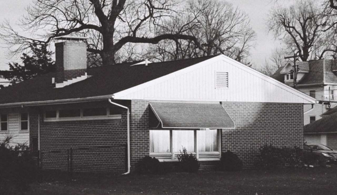 Black and white photo of a mid-century brick ranch house with a gabled white siding end, brick chimney