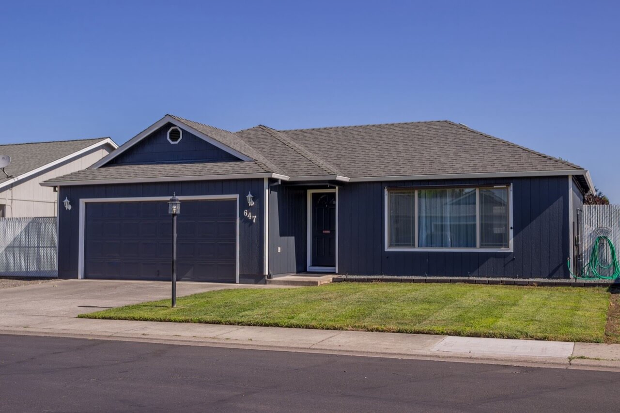 A single-story navy-blue ranch house with a two-car garage, tidy green lawn, and concrete driveway sits on a quiet suburban street under a clear blue sky.