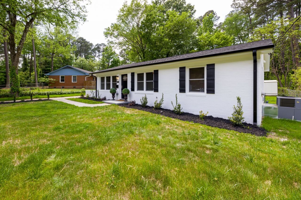 A freshly renovated white brick single-story ranch house with black shutters, new landscaping mulch beds, and a wide green lawn in a leafy suburban neighborhood.