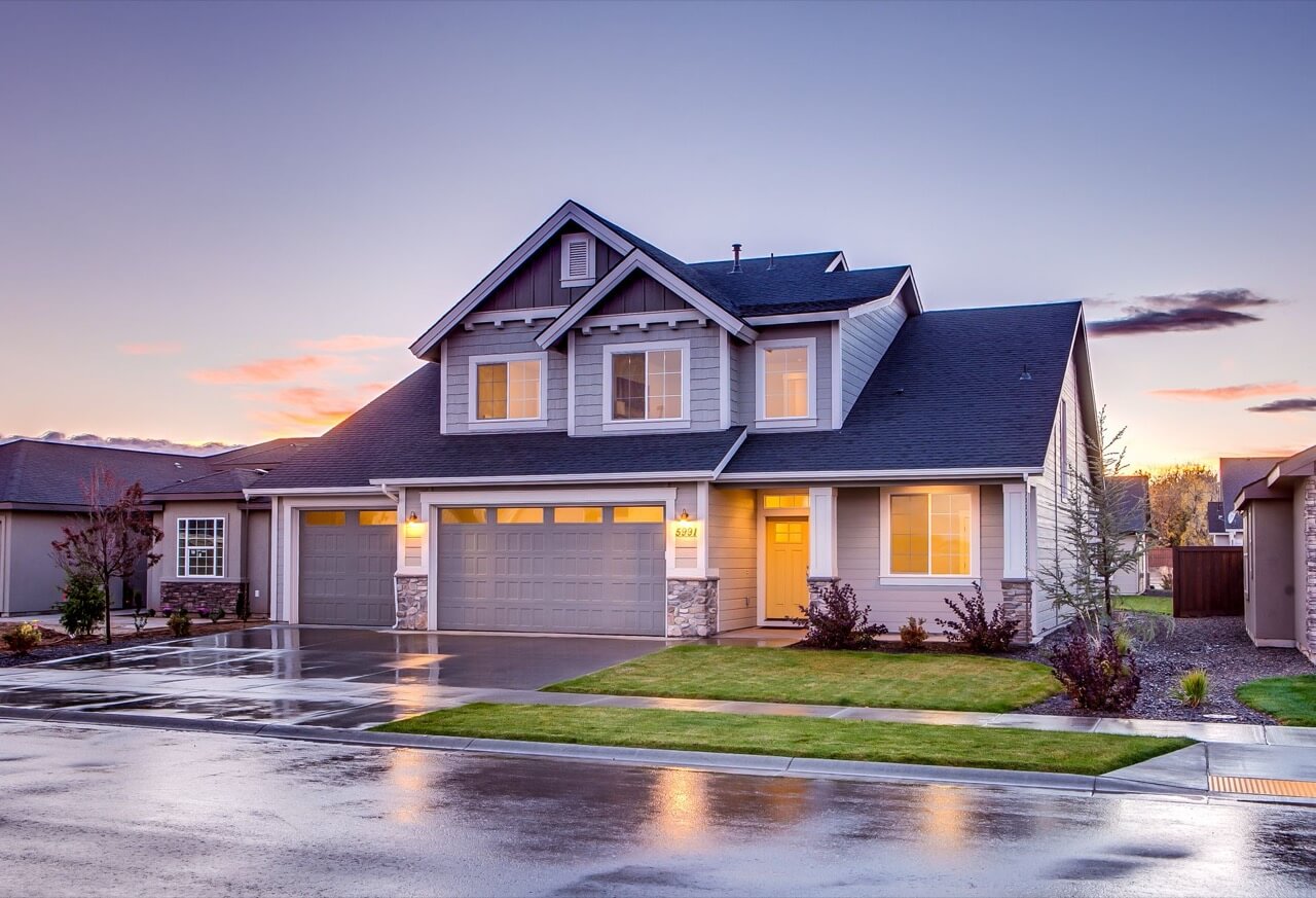 A blue-gray craftsman-style suburban house with a three-car garage and warmly lit windows glows at dusk