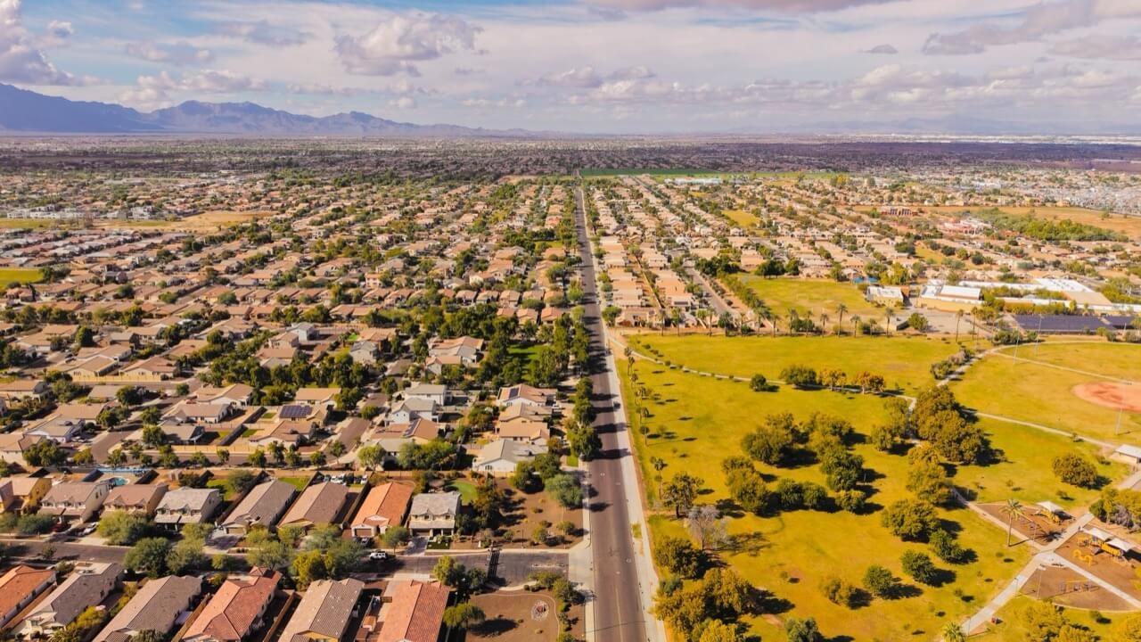 Aerial view of a sprawling desert suburban neighborhood with tan tile-roofed homes along a straight road beside a green