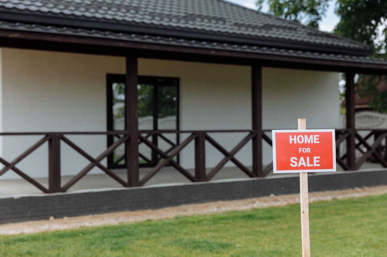 A red home for sale yard sign on a wooden stake stands on a green lawn in front of a blurred white house with a dark tiled